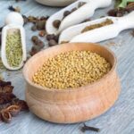 Mustard seeds in a red clay bowl surrounded by an assortment of spices on a wooden table.