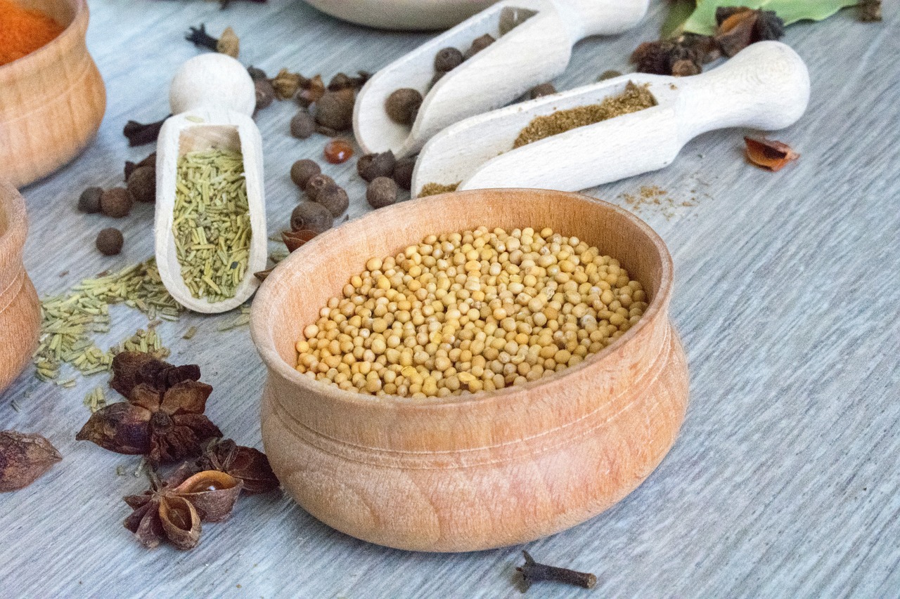 Mustard seeds in a red clay bowl surrounded by an assortment of spices on a wooden table.